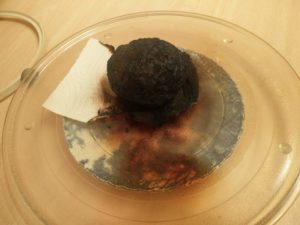 The potato out of the microwave oven, showing the blackened remains of itself and the paper